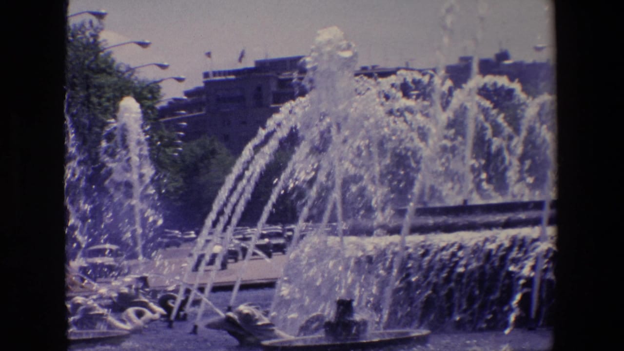 1967 Madrid 8mm frame showing a sunlit outdoor fountain with multiple water jets, cascading tiers, distant buildings, streetlights, and cars partly visible behind the spray.