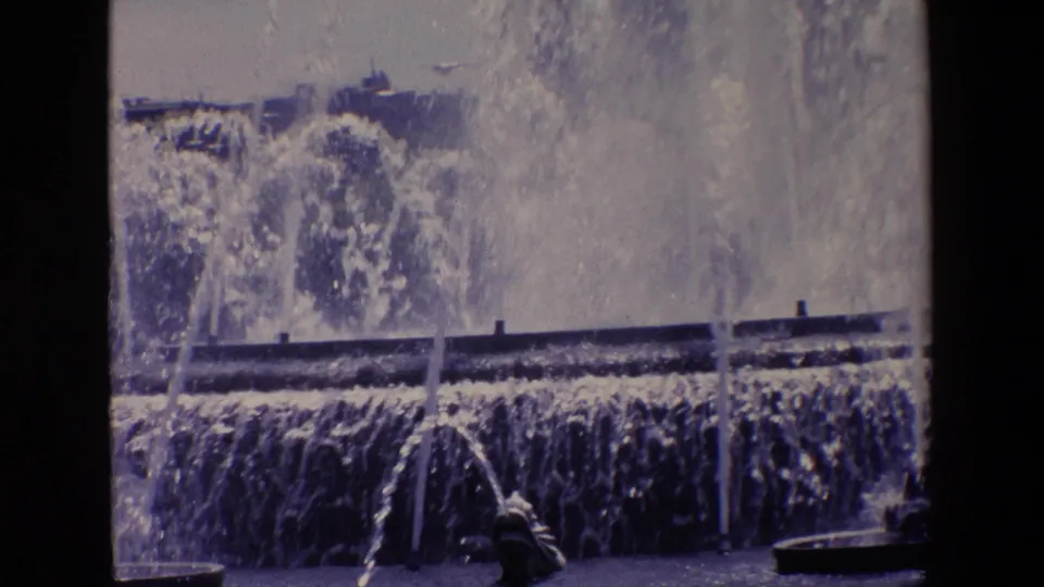 1967 Madrid 8mm frame focused on dense fountain spray and a dark horizontal basin edge, with urban buildings and streetlights softened in the background.