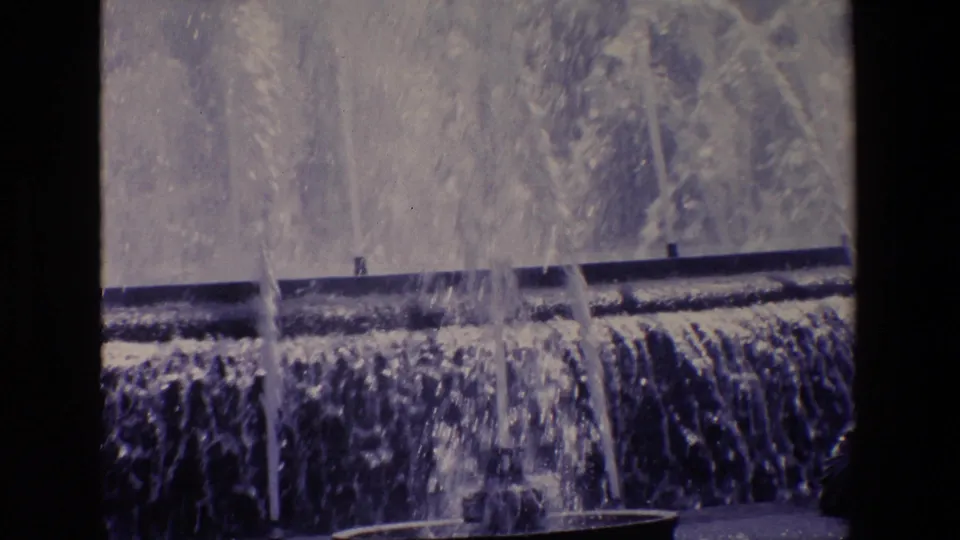 1967 Madrid 8mm frame showing fountain water cascading over a broad tier, with tall spray columns and soft blue-tinted film texture dominating the view.