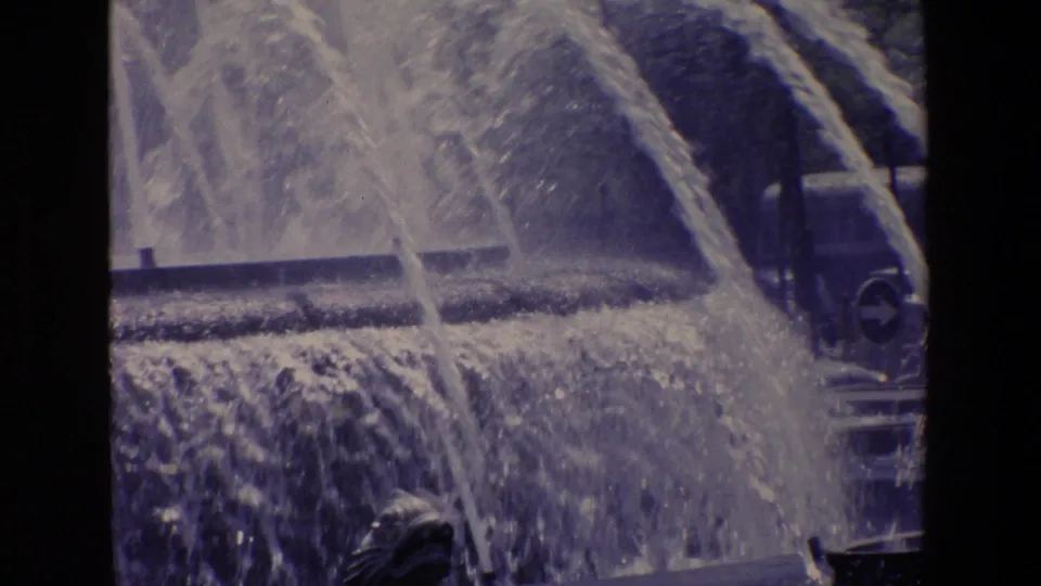 1967 Madrid 8mm frame with crossing fountain jets, cascading water, a dark basin edge, and a partially visible bus or vehicle at the right side of the plaza background.