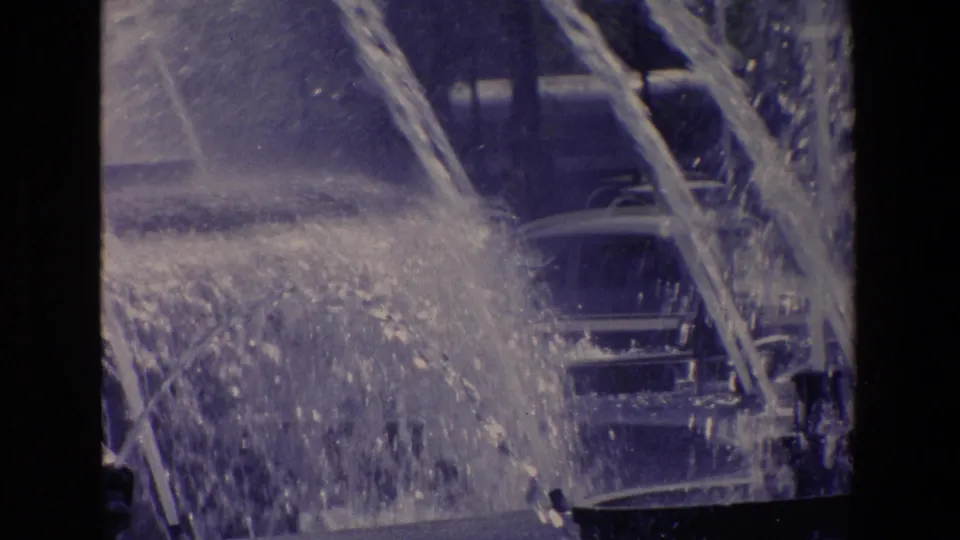 1967 Madrid home-movie frame with arcing fountain water partly covering classic cars in the background.