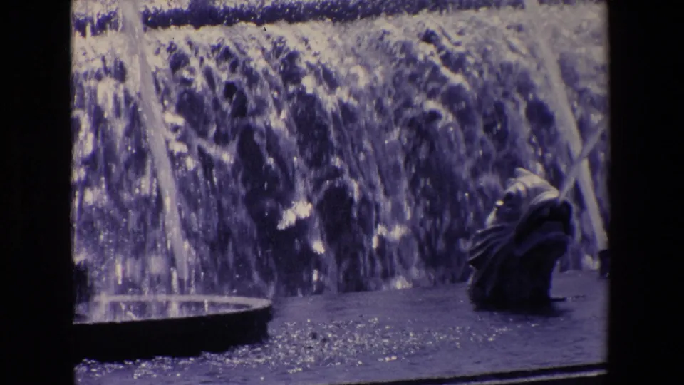 Close view of foaming fountain water and a small sculptural spout, with city traffic mostly hidden beyond the spray.