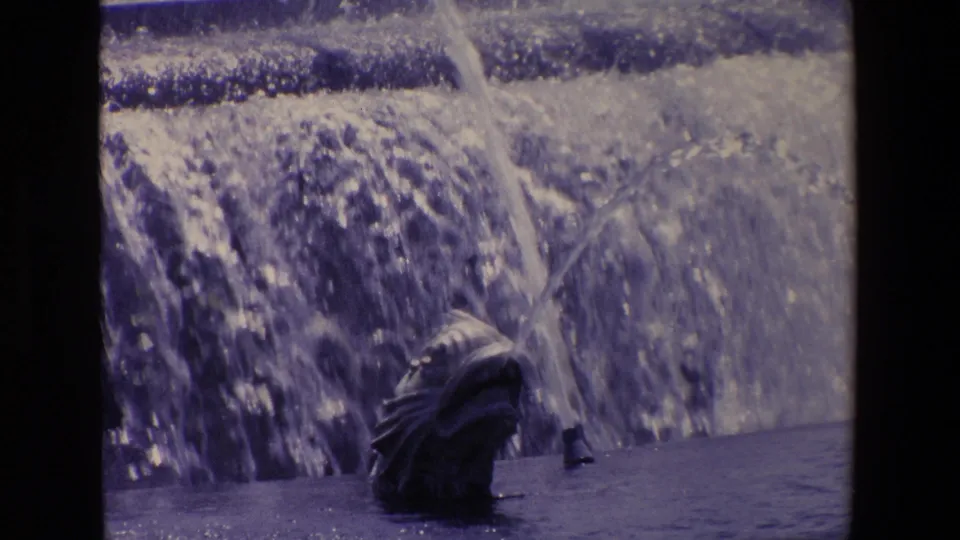 Fish-shaped fountain spout sits near the lower frame as water arcs across a blurred Madrid street background.