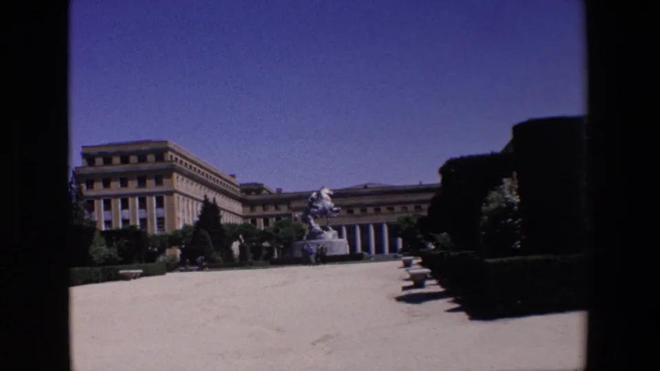 Wide 1967 Madrid home-movie view across a paved formal courtyard toward a grand building, clipped hedges, and a central equestrian statue with small distant visitors.