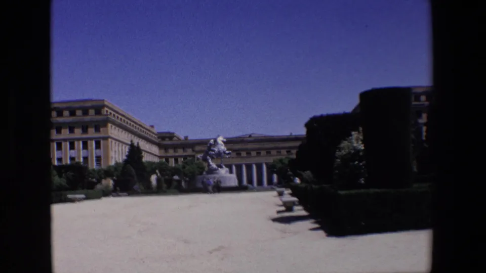 Wide 1967 Madrid home-movie frame showing the formal courtyard from a similar angle, with the building facade, equestrian statue, hedges, and open sky visible.