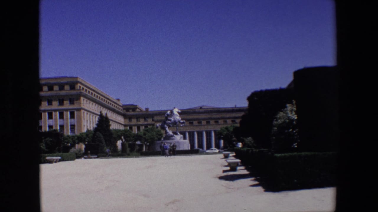 Wide view of a paved square with an equestrian statue, columned building, hedges, trees, and dark film borders.
