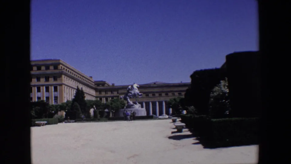 1967 Madrid home-movie frame showing a sunlit formal square with a large equestrian statue before a long neoclassical building, with hedges and trees around the plaza.