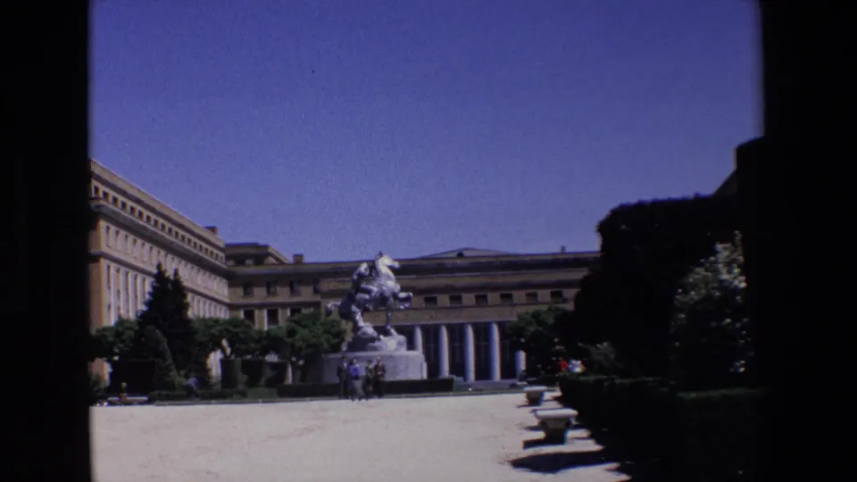 1967 Madrid exterior view with the equestrian statue centered near a columned building, framed by manicured hedges, benches, trees, and the black edges of the film scan.