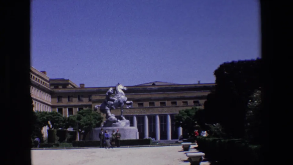1967 Madrid home-movie frame showing visitors near the base of a large equestrian monument, with a formal building facade, columns, hedges, and trees in the background.