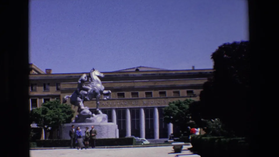 1967 Madrid frame with the equestrian monument in front of an inscribed neoclassical facade, several pedestrians at the base, clipped hedges, trees, and a bright open plaza.