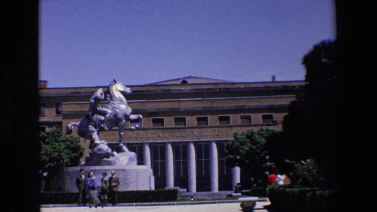 1967 Madrid home-movie frame showing an equestrian statue on a pedestal in front of the Facultad de Medicina building, with columns, trees, and small groups of pedestrians visible.