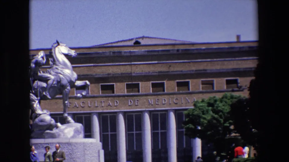 1967 Madrid frame with the silver-toned horse-and-rider statue partly centered before the Facultad de Medicina entrance, showing engraved lettering, columns, windows, greenery, and distant pedestrians.