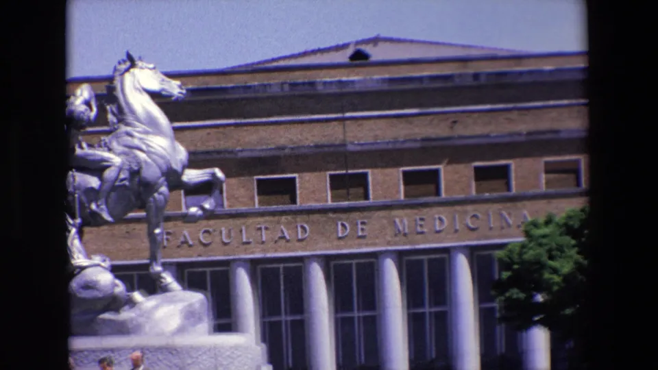 1967 Madrid home-movie frame tightly cropped on the equestrian statue and the Facultad de Medicina facade, with the stone pedestal, upper windows, entrance columns, and a few people at the lower edge.