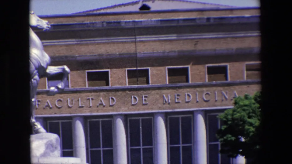 1967 Madrid frame showing the Facultad de Medicina lettering across the facade, entrance columns and windows below, tree foliage at right, and a partial view of the equestrian statue at left.