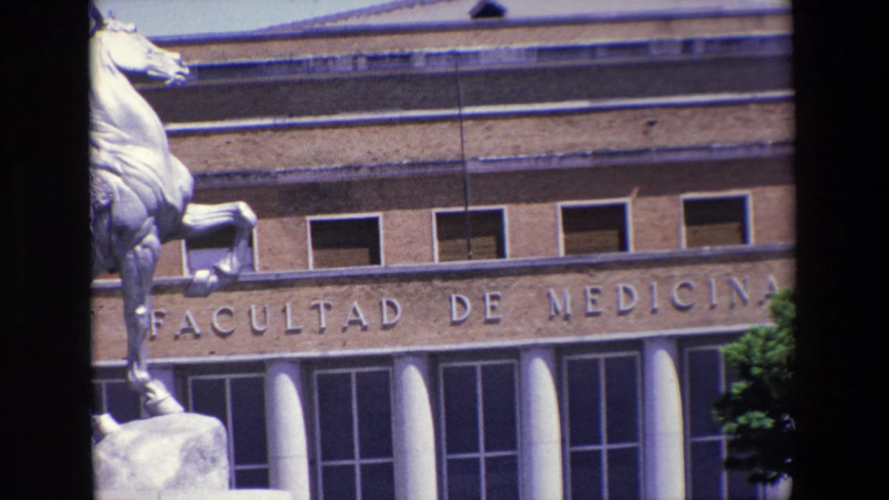 1967 Madrid home-movie frame showing the Facultad de Medicina facade with engraved lettering, upper windows, white columns, and part of an equestrian statue at the left edge.