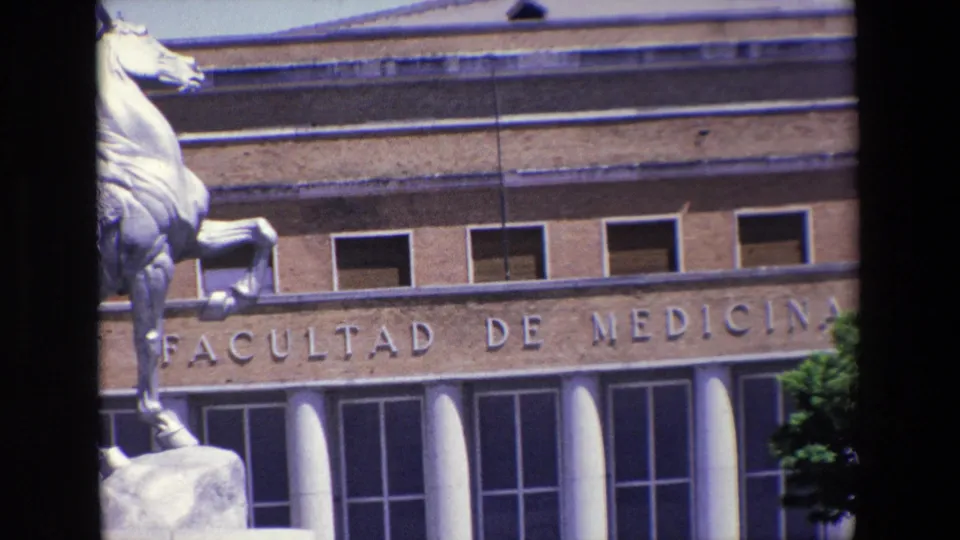 1967 Madrid home-movie frame showing the Facultad de Medicina facade with engraved lettering, upper windows, white columns, and part of an equestrian statue at the left edge.