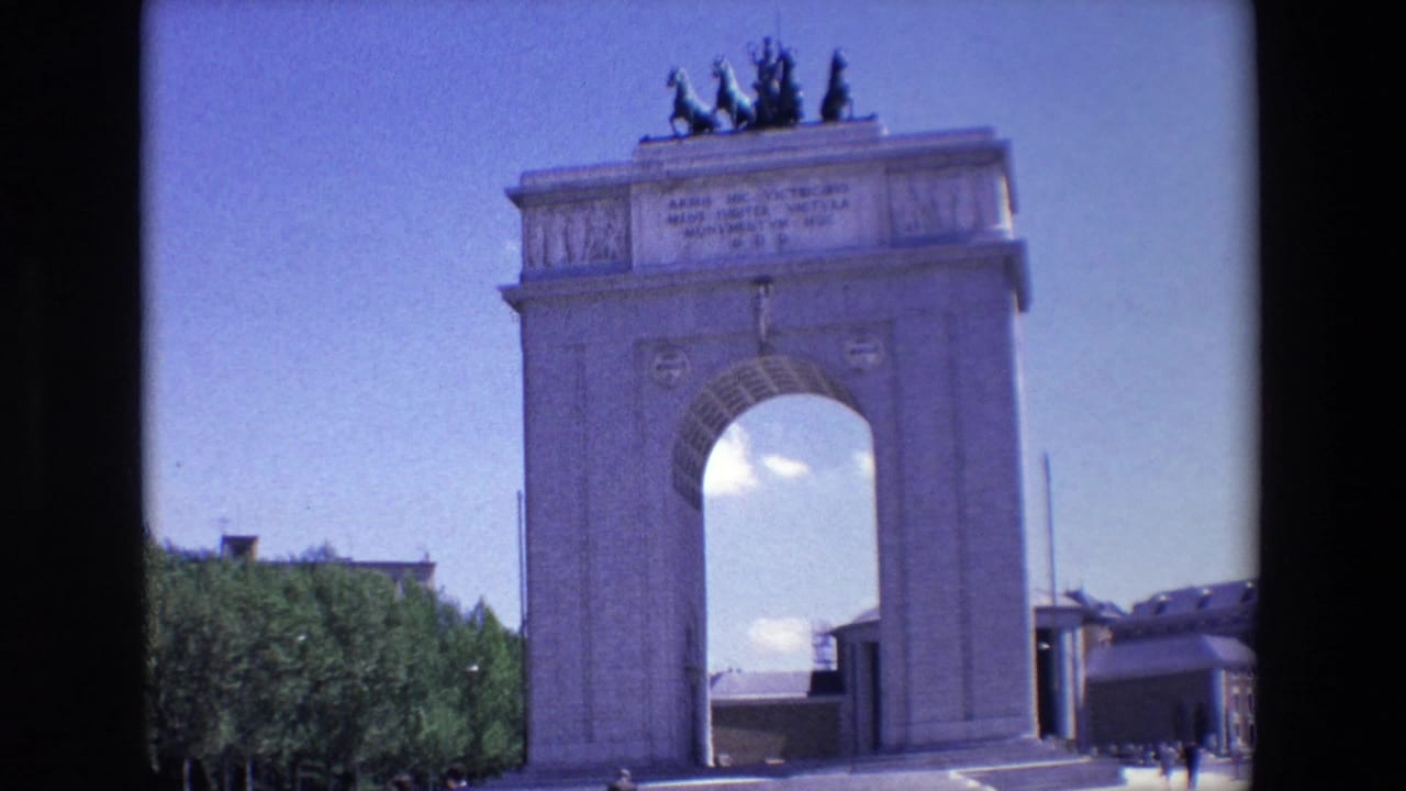 1967 Madrid 8mm frame showing the Monumento a los Caidos por Espana with a central arch, rooftop statues, trees, and distant visitors under a blue sky.