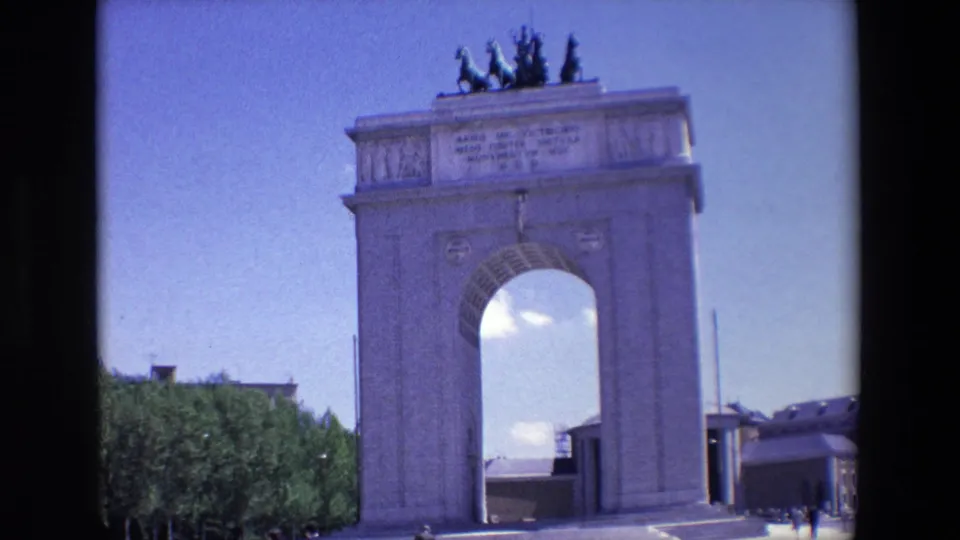 1967 Madrid 8mm frame showing the Monumento a los Caidos por Espana with a central arch, rooftop statues, trees, and distant visitors under a blue sky.