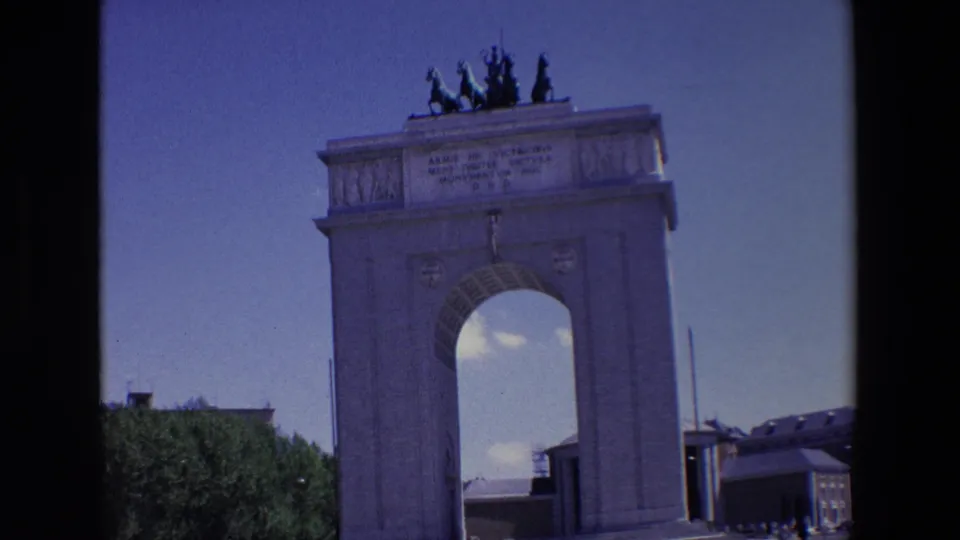 1967 Madrid home-movie frame of the monument from a slightly closer ground-level view, showing the arched opening, upper inscription, rooftop statuary, and tiny figures below.