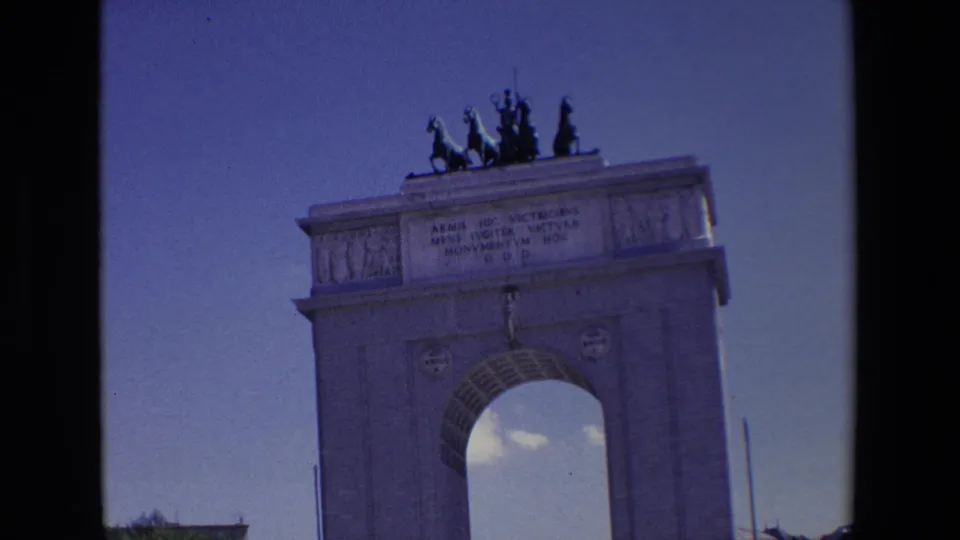 1967 Madrid 8mm frame focused on the upper portion of the monument, with the inscription, rooftop figures, relief panels, and part of the arch opening visible.
