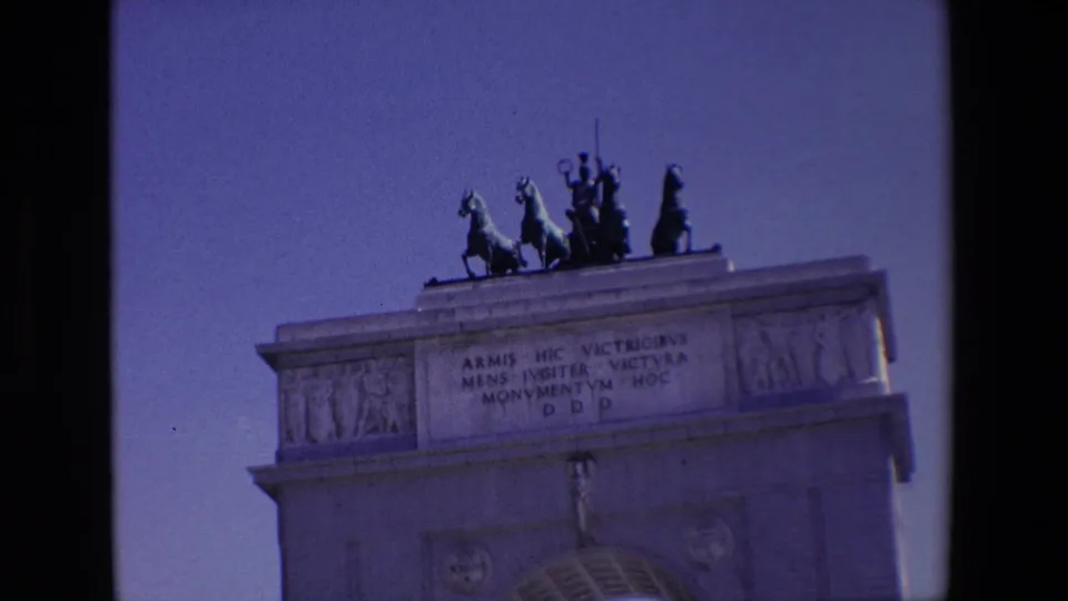 1967 Madrid home-movie frame showing the monument's rooftop sculptural group and Latin inscription, with the top of the arch cropped low in the frame.