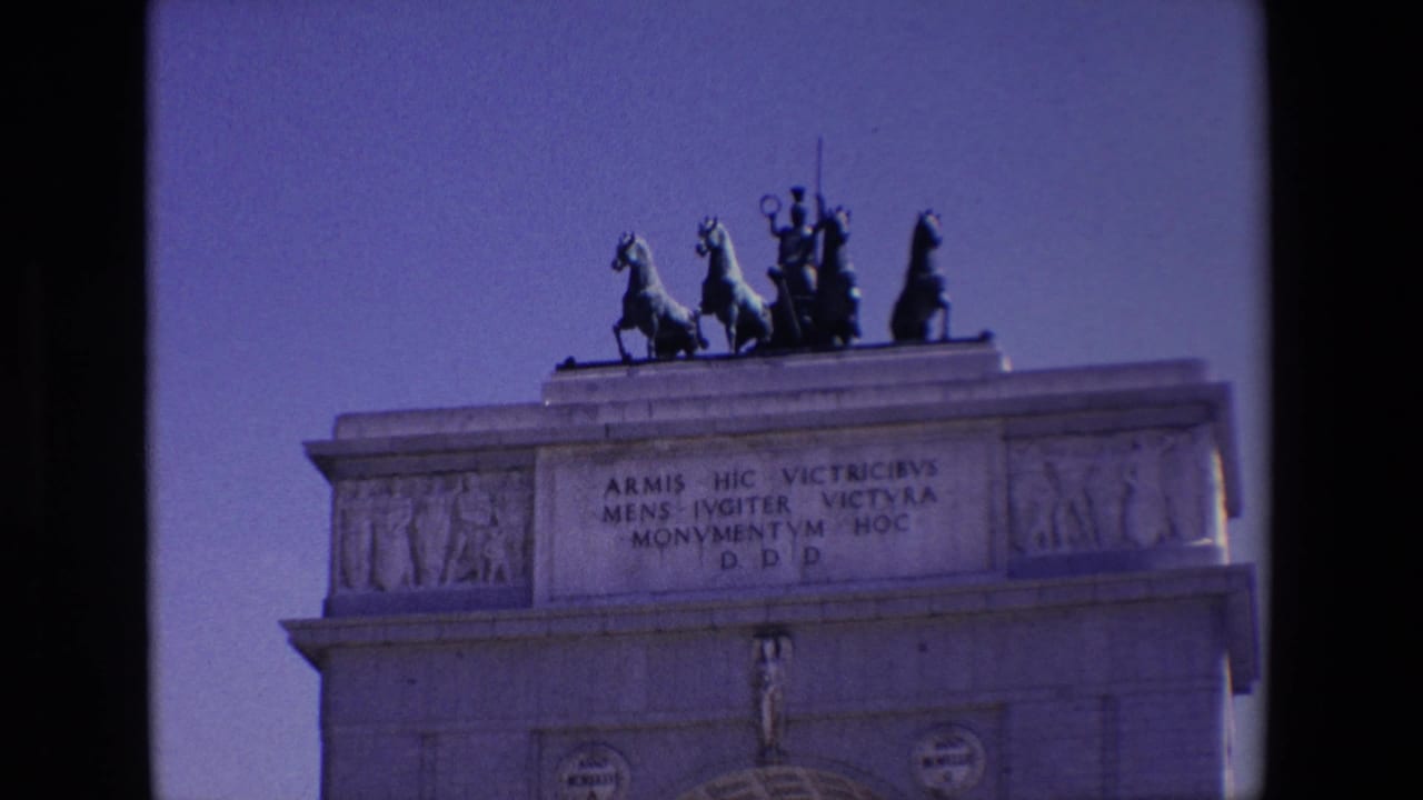 Upper portion of a stone triumphal arch with a quadriga sculpture against blue sky.
