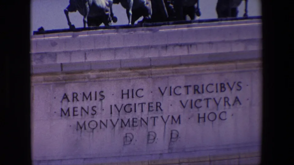 Zoomed 8mm frame from Madrid in 1967 showing carved Latin text on the Victory Arch facade, with part of the rooftop quadriga visible above.