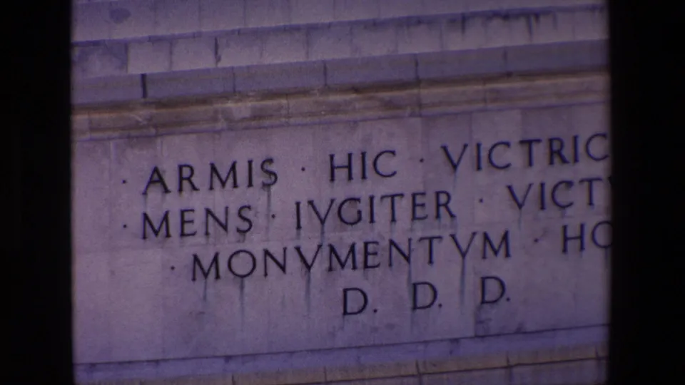 1967 Madrid home-movie frame showing a close-up of a stone monument inscription with Latin lettering, partially obscured by dark frame edges.