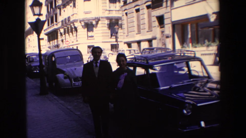 1967 Madrid home-movie frame showing a man in a dark suit and a woman in dark clothing with a light scarf walking along a cobblestone street beside parked vintage cars.