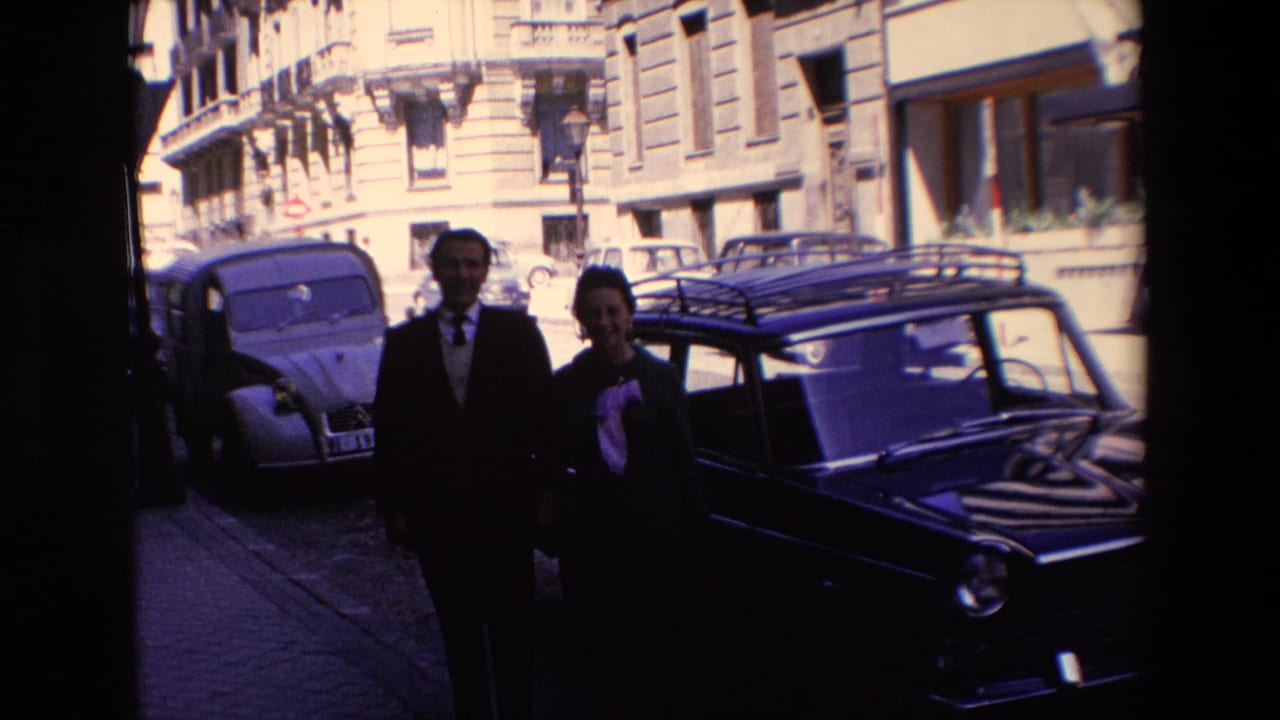 In Madrid, Spain, according to the supplied 1967 metadata, a suited man and a woman in a dark coat stand near parked compact cars on a bright city street.