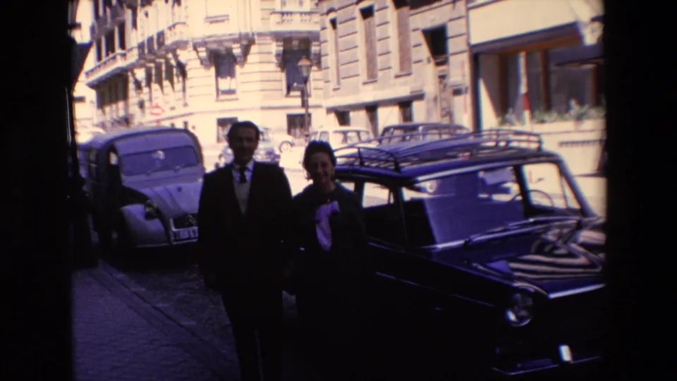 In Madrid, Spain, according to the supplied 1967 metadata, a suited man and a woman in a dark coat stand near parked compact cars on a bright city street.