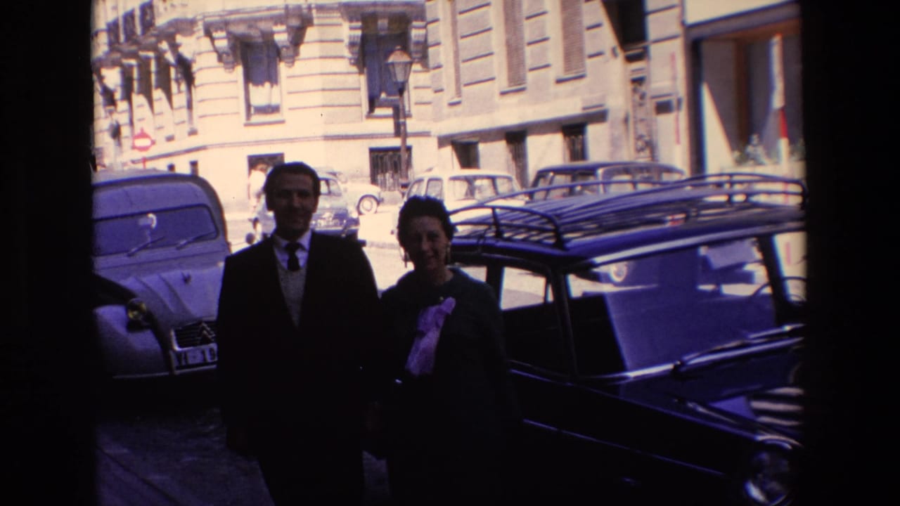 Couple closer to the camera with parked cars and stone-fronted buildings behind them.