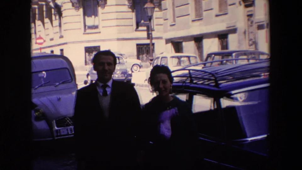 The couple remains posed in partial shadow on the street, with a compact car, a dark roof-rack vehicle, apartment windows, and bright pavement behind them.