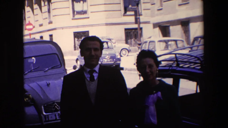 A closer 8mm frame shows the smiling couple in Madrid, based on the metadata, standing near parked European cars with an apartment building and street sign behind them.