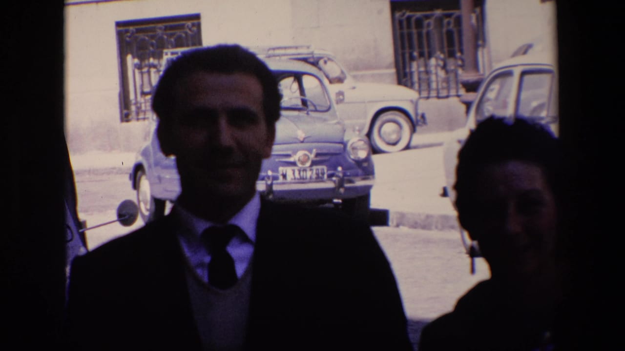 Suited man stands close to the camera with a woman partly visible at the edge.