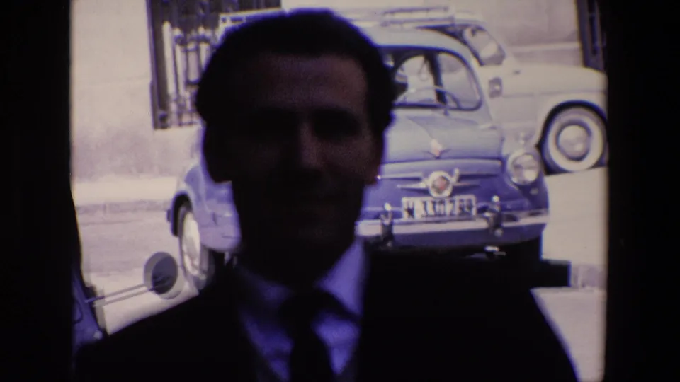 The camera moves closer to the suited man, leaving his face softly rendered in shadow while a blue compact car, another parked car, and a building wall remain visible in the sunlit background.