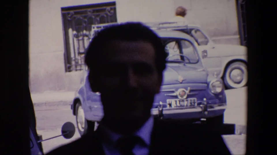 A soft, dark close view shows the suited man centered in front of a bright street scene with a blue compact car, another small car, and a barred window on the building behind him.