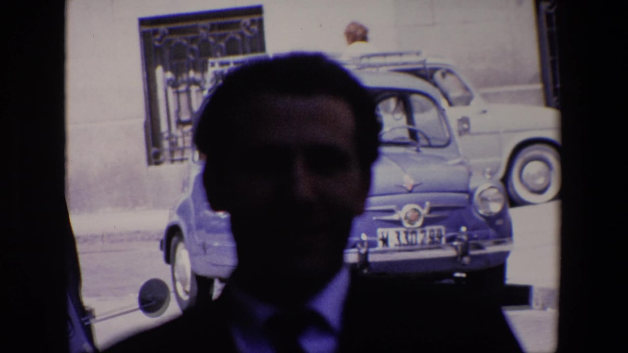 Silhouetted man in a suit stands close to camera with a blue compact car and barred window behind him.