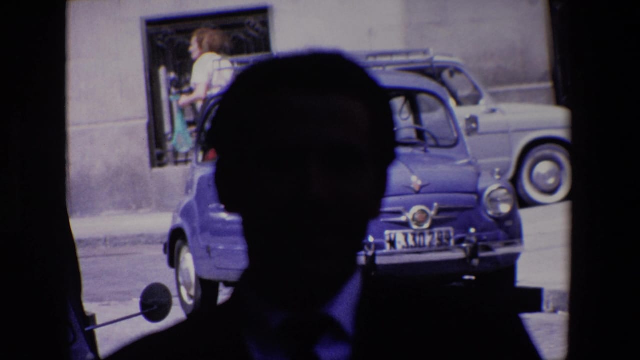 Dark foreground silhouette overlaps a parked blue compact car as a woman walks near the building wall.