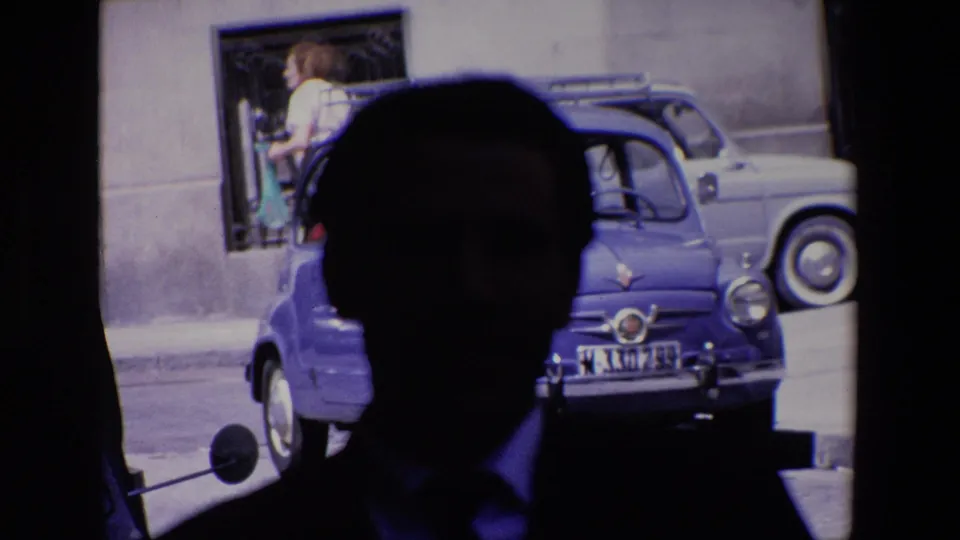 Madrid, Spain, 1967 frame with a shadowed male figure close to the lens, a blue compact car parked behind him, and a woman in light clothing passing along the street near a barred window.