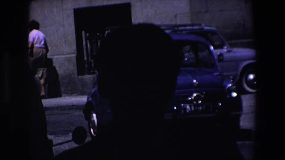 Madrid, Spain, 1967 8mm frame showing a blue compact car in heavy shadow beside a building entrance, with a man at the left edge walking near a dark suitcase or bag.