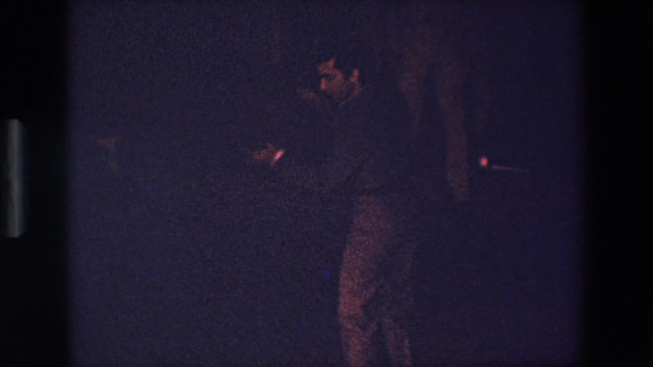 Adult performer in dark clothing stands in profile against a dim background.