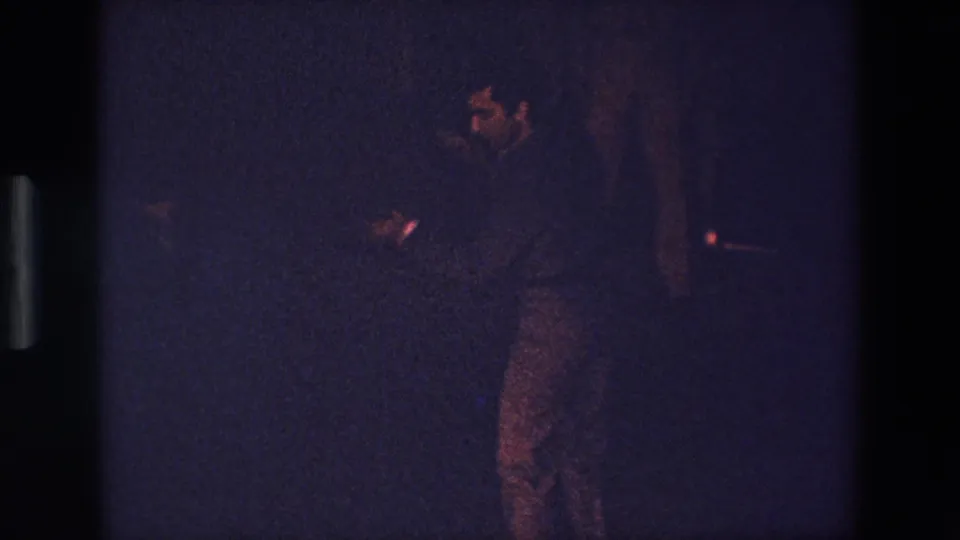 A grainy 1967 Palermo home-movie frame shows an adult performer in dark clothing seen in profile, with hands raised or extended in a dim indoor setting.
