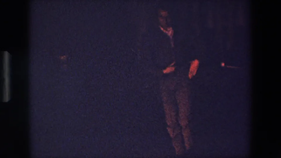 A low-exposure 1967 Palermo 8mm frame shows an adult performer facing the camera area, with reddish hands and face visible against a dark indoor background.