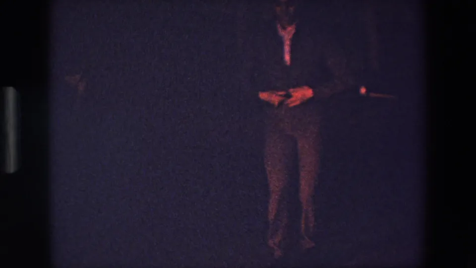 A grainy 1967 Palermo home-movie frame shows an adult performer standing with hands near the waist, partly silhouetted against a dark purple-blue indoor background.