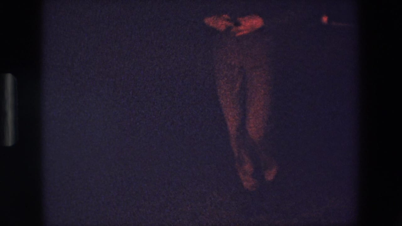 1967 Palermo 8mm frame showing a standing male dancer partially visible in low light, with his legs and hands catching reddish illumination against a dark background.