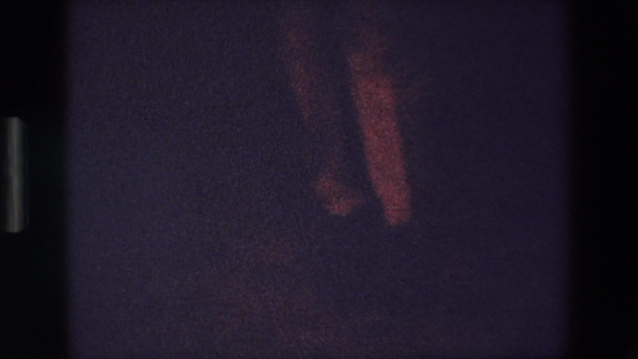 Dim frame with a dancer's lower body and one arm faintly visible against darkness.