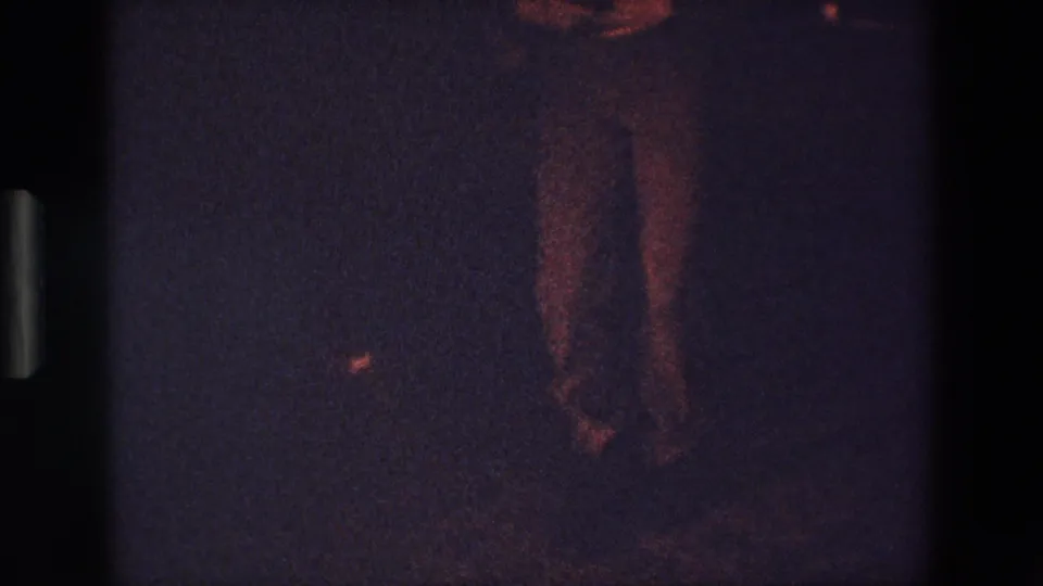 1967 Palermo 8mm frame showing the dancer's lower torso and legs more clearly, with reddish light on dark trousers and a shadowed floor area.