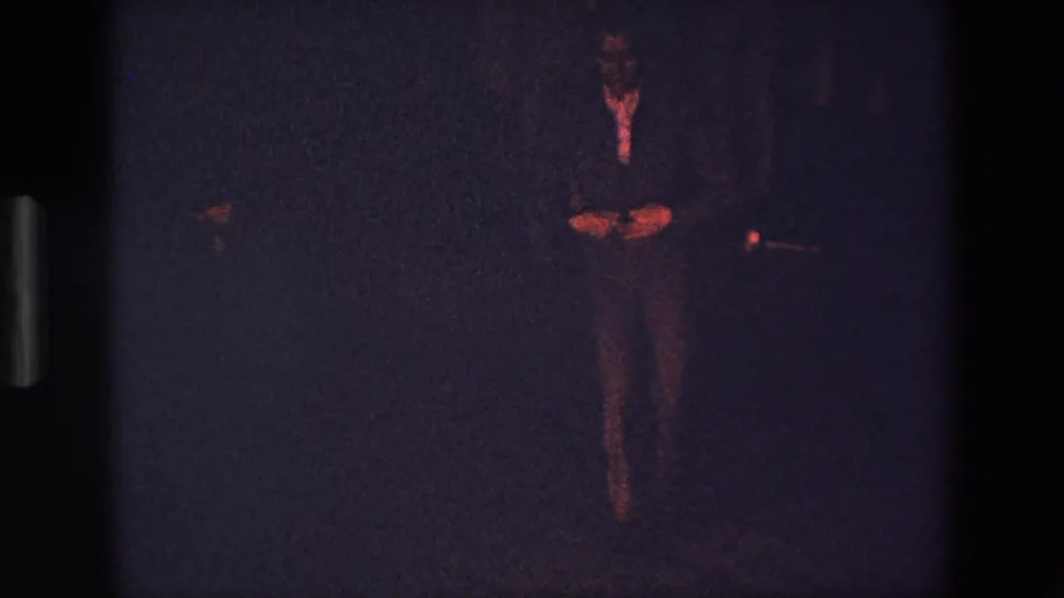 1967 Palermo 8mm frame showing the male dancer as a small full-body figure, with hands and legs highlighted by reddish light against a mostly dark background.