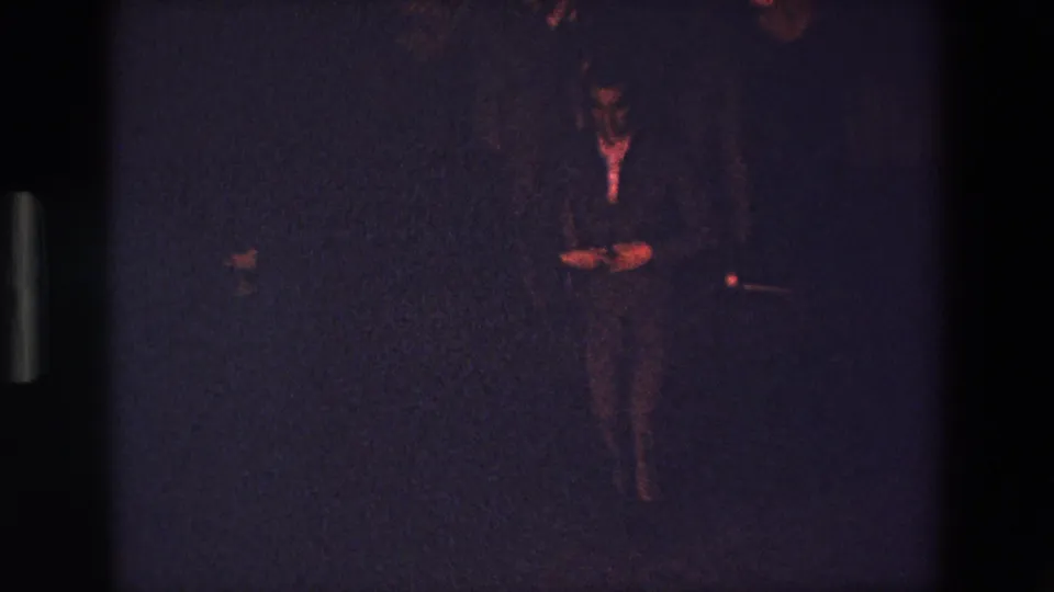 A dark-clad figure appears in a grainy, underexposed 1967 Palermo home-movie frame with reddish highlights and little visible background detail.